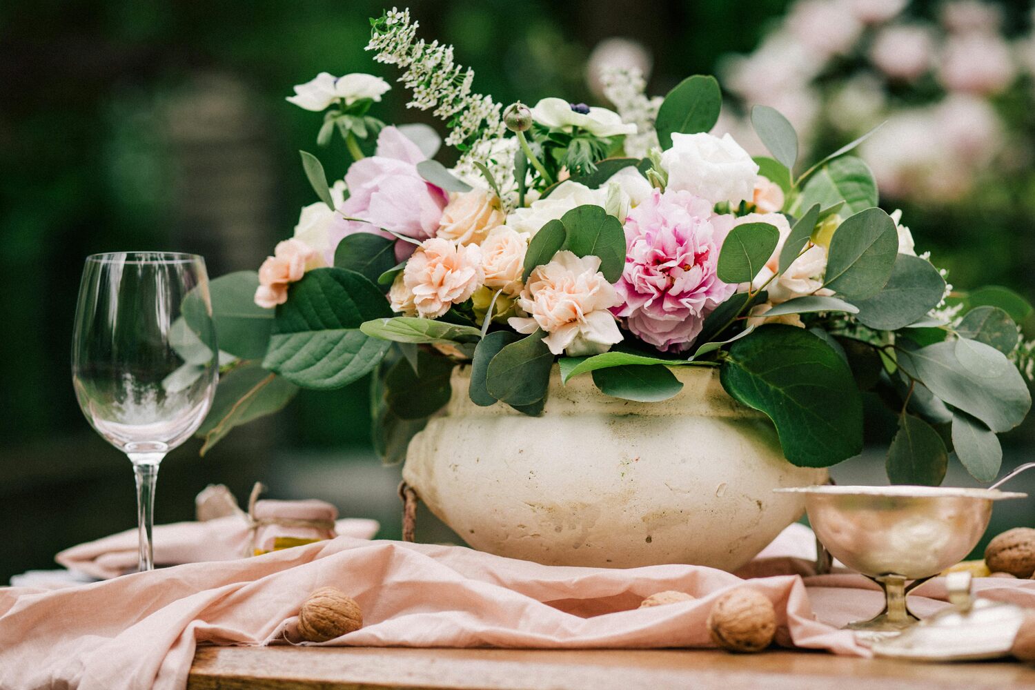 A floral arrangement featuring pink peonies and other flowers in a ceramic vase with green leaves, set on a table with delicate linens.